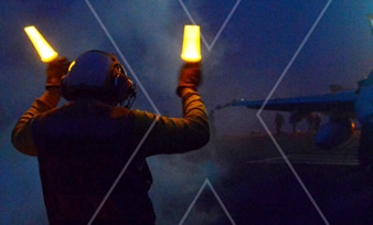 NavalX| decorative image of an aircraft marshaller at night fuiding a plane landing on a naval cruiser